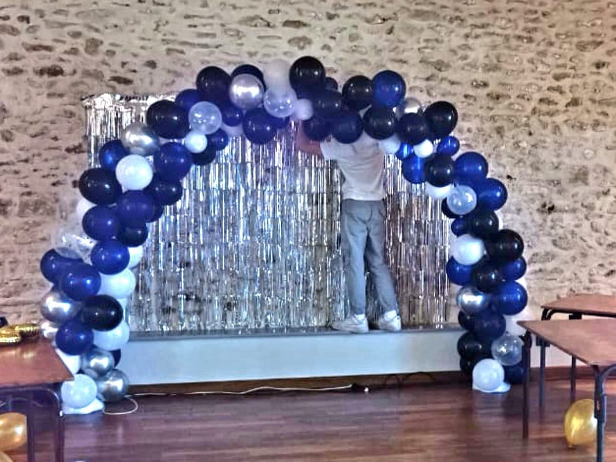 Arche de ballons bleus, blancs et argentés montée devant un rideau argenté dans une salle